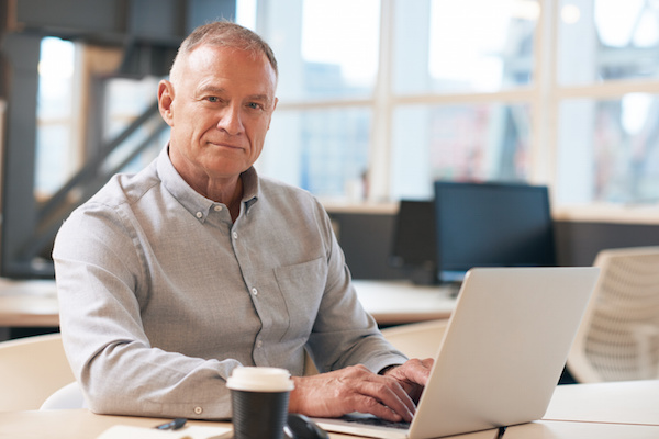 Portrait of a confident mature businessman wearing casual clothes sitting at a desk in a modern office working on a laptop
